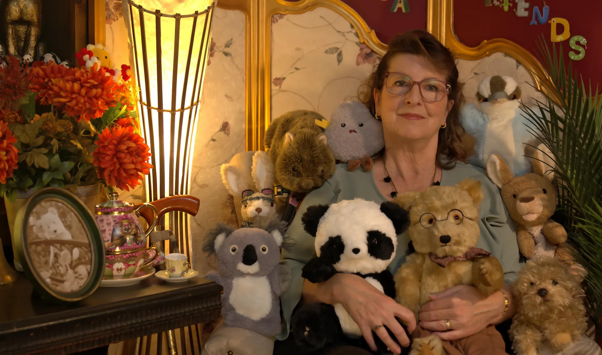 Miss Lisa seated among her soft toy friends, beside a lit lamp and a vase of autumn flowers